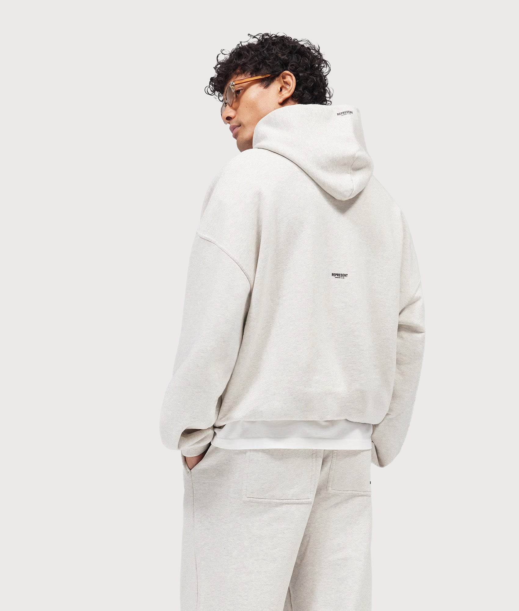 Represent Oversized Micro Owners Club Hoodie in Cream Marl at EQVVS Menswear. Angled model back shot.