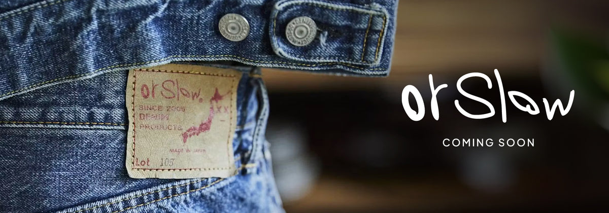 OrSlow denim jeans label on a close-up of blue denim, with the OrSlow logo and “Coming Soon” text on a dark, blurred background.